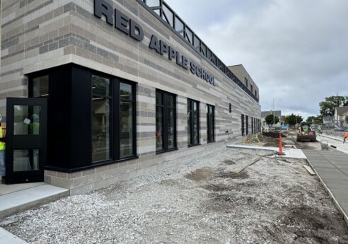 Modern brick school building labeled Red Apple School under construction, with large windows, gravel lot, sidewalk, and a cloudy sky.