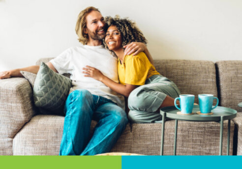 A couple sits on a beige sofa, smiling and hugging, wearing casual clothes, with a small round table holding teal mugs nearby in a bright living room.