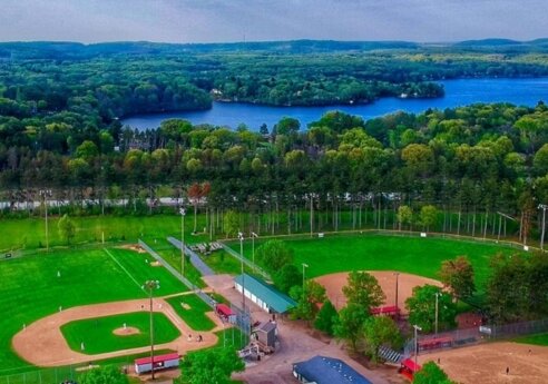 Aerial view of a baseball complex with multiple fields, surrounded by trees, a blue lake beyond, and forested hills on the horizon.