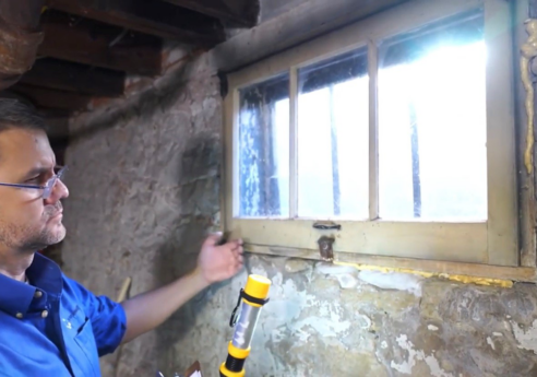 Person in a blue shirt and glasses gestures toward a small latch on an old basement window, holding a yellow flashlight; peeling walls and exposed pipes surround.