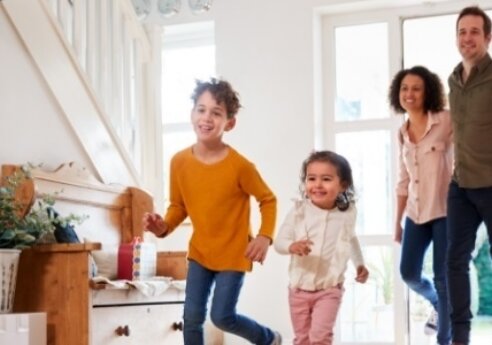 A family of four, two adults and two children, runs through a bright living room with a staircase, plants, and large windows, smiling.