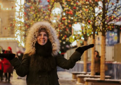 A person in a dark parka with a fluffy fur-lined hood stands outdoors at night, arms outstretched, smiling among festive holiday lights.