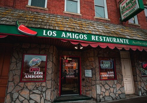 Front of Los Amigos Restaurante, a stone storefront with a green awning, chili pepper sign, and neon OPEN windows for authentic Mexican cuisine.