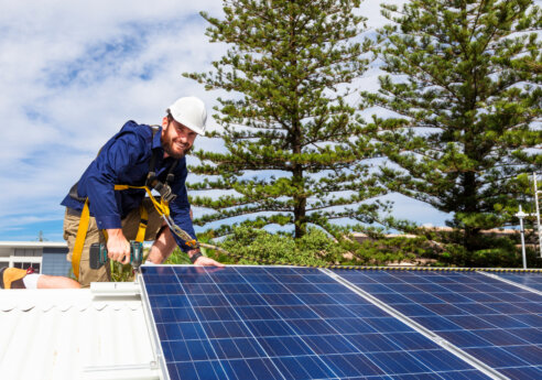 Person wearing a white hard hat and blue work shirt, on a roof installing blue solar panels with a drill, secured by a safety harness, with trees and a blue sky behind.
