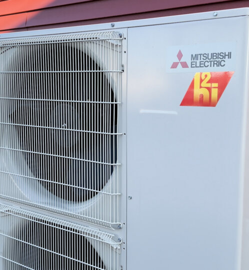 Outdoor Mitsubishi Electric heat pump/condenser unit mounted outside a red siding house, with a large protective grille over two fans and a visible logo.