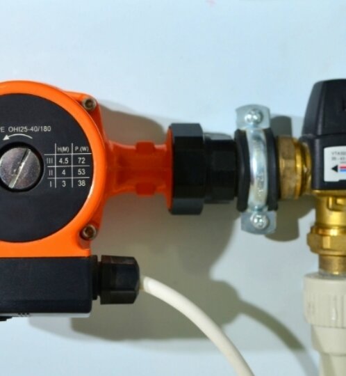 Orange circulator pump with black dial, mounted on piping connected to brass valve fittings; a white cable trails beneath.