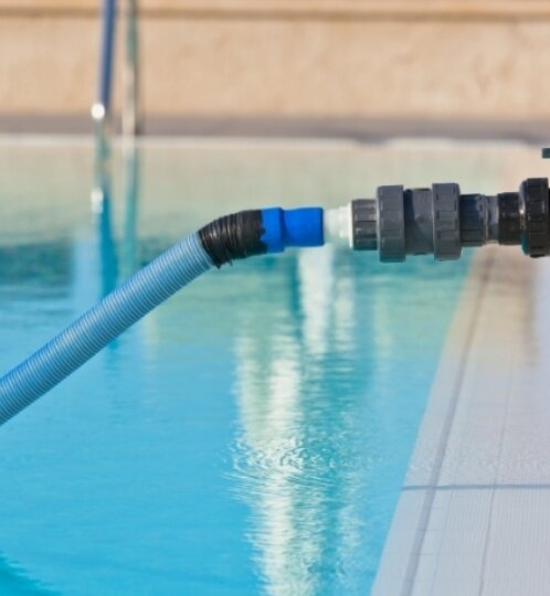 Blue flexible pool hose attached to a pump at the pool edge, turquoise water, and a metal ladder in the background.