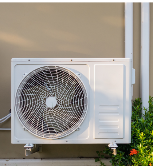 White outdoor air conditioner condenser mounted on a beige wall, featuring a circular fan grille and a vent panel, with green plants nearby.