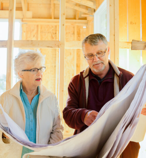 Two adults with glasses examine a large blueprint inside a partially constructed wooden-framed room.