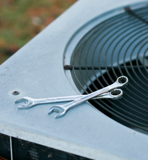 Two silver wrenches resting on the metal housing of an outdoor air conditioner condenser, near the circular fan grille.