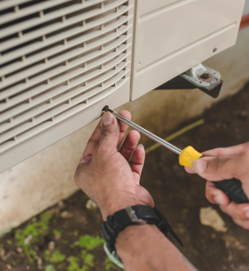 Close-up of hands using a screwdriver to tighten a screw on an outdoor beige HVAC condenser unit with a vented cover and rusted bracket.