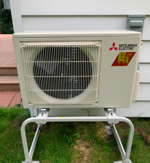 Beige Mitsubishi Electric outdoor unit on a white metal stand, circular fan behind a grille, beside house siding and a wooden deck.