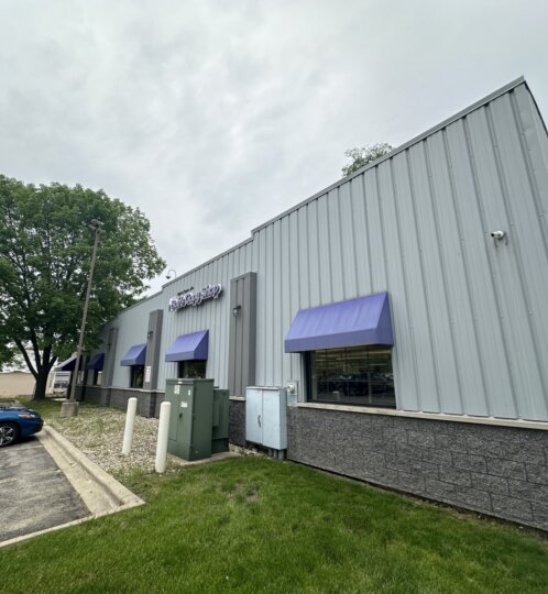 Angled view of a gray metal commercial building with purple awnings, a blue car in the parking lot, and a large tree beside the lot under a cloudy sky.