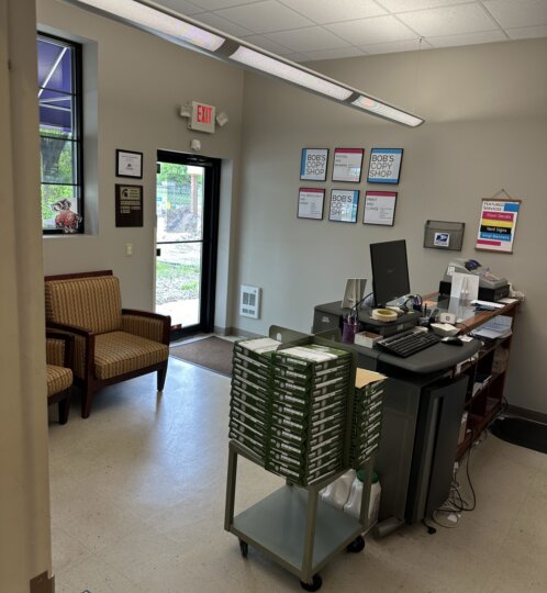 Small shop interior with a front desk computer, a cart stacked with green boxes, two upholstered chairs, an exit door, and wall posters.