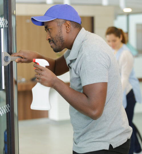 A person wearing a blue cap cleans a glass door with a spray bottle in a bright indoor lobby; another person is blurred in the background.