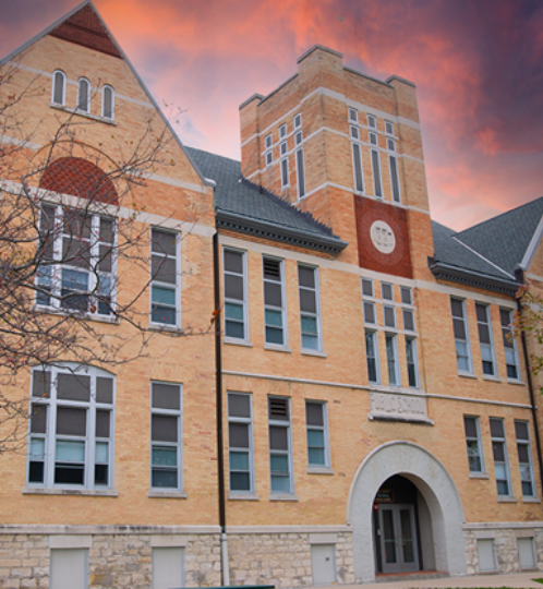 Brick campus building with an arched entry, tall clock tower, and many windows; stone base, leafless trees, and a pink-orange sunset sky.