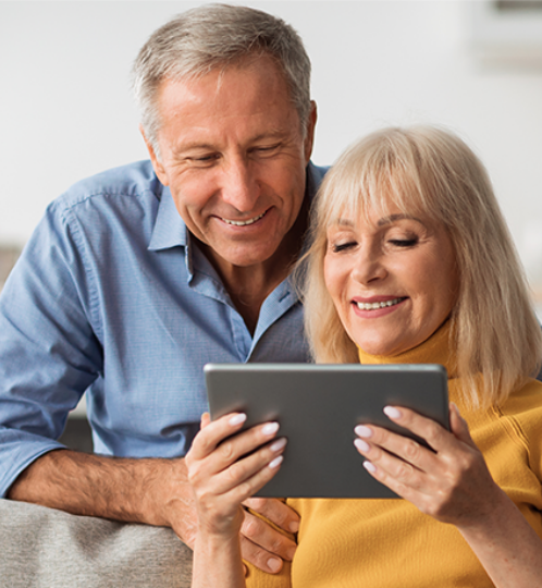 Two adults sit close on a couch, smiling at a tablet; one has an arm around the other in a warm, bright home setting.