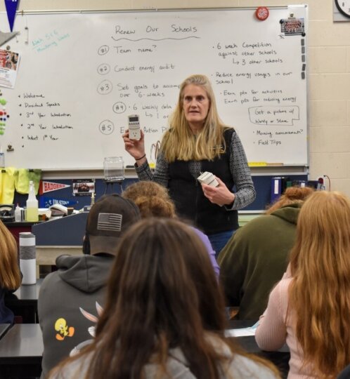 Person with long blonde hair stands at the front of a classroom, presenting to students; a whiteboard with notes sits behind.