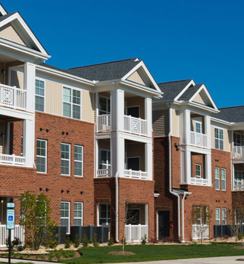 Row of brick-and-beige townhouse-style apartment buildings with white balconies, peaked roofs, and a neatly manicured lawn under a bright blue sky.