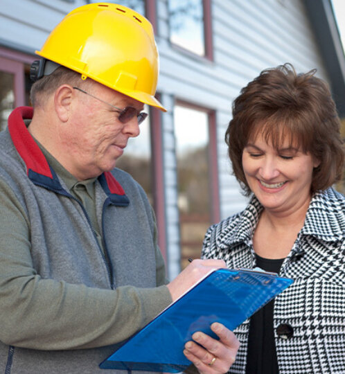 Two adults outside a blue building; one in a yellow hard hat and gray vest shows a clipboard to the other, who smiles in a checkered jacket.