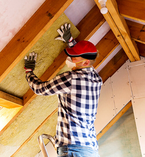 Person in a plaid shirt, mask, and gloves stands on a ladder in an attic, smoothing insulation panels onto a sloped wall between wooden beams.