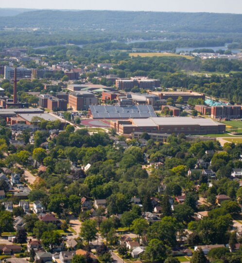 Aerial view of a tree-filled suburb with a campus of brick buildings and a track, surrounded by houses and distant hills.