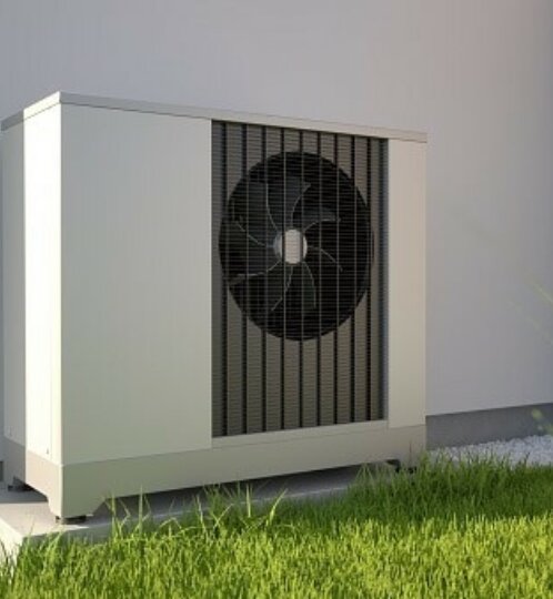 Outdoor air conditioner condenser unit on a small concrete pad beside a white wall, with a visible fan behind a grille, surrounded by gravel and green grass.
