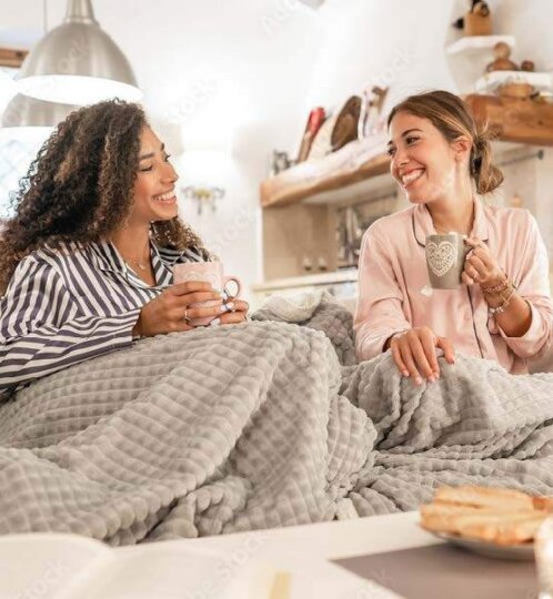 Two people sit on a couch under a gray quilted blanket, smiling at each other while holding mugs in a cozy, sunlit living room.