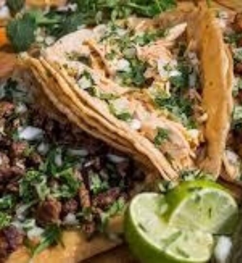 Plate of soft corn tacos filled with meat, topped with chopped cilantro and onions, with lime wedges and salsa on a wooden table.