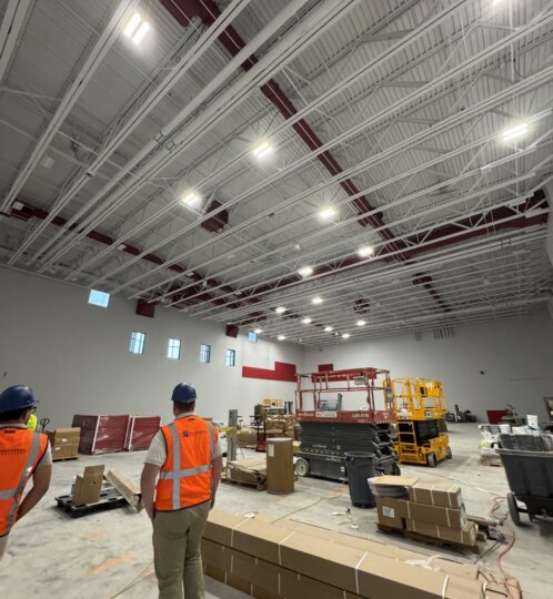 Two construction workers in orange vests and blue hard hats walk through a large, unfinished warehouse with exposed ceilings and scattered boxes.