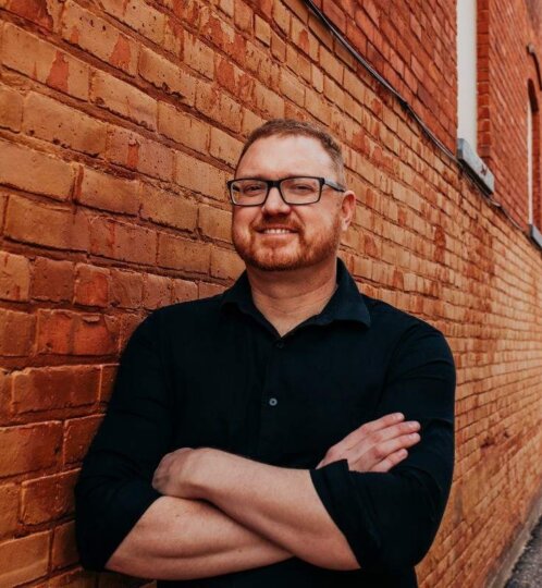 Person with short hair and beard, wearing a black shirt and glasses, arms crossed, smiling, standing against a red brick wall.