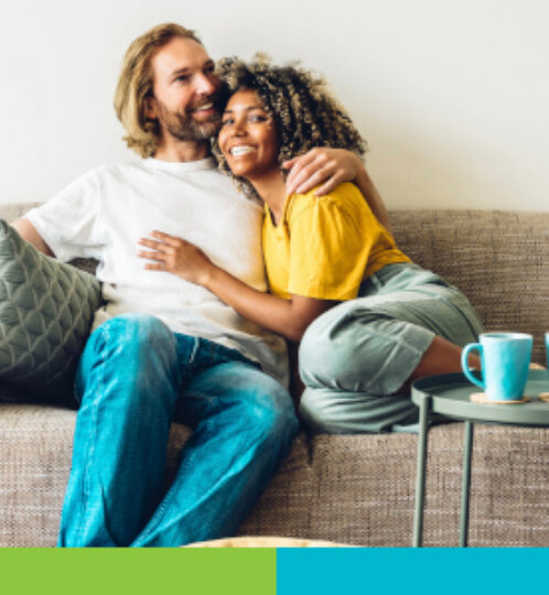 A couple sits on a beige sofa, smiling and hugging, wearing casual clothes, with a small round table holding teal mugs nearby in a bright living room.