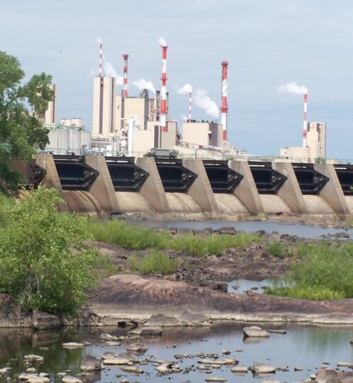 Industrial power plant with red-and-white smokestacks behind a concrete dam; rocks and green vegetation line a calm river in the foreground.