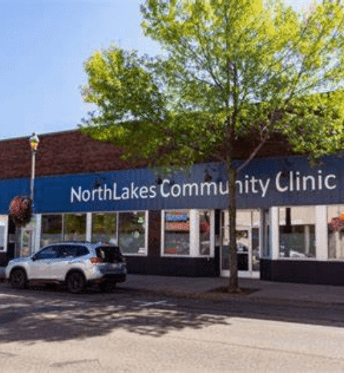 Front view of NorthLakes Community Clinic storefront with a blue awning, large windows, a tree in front, and parked cars on a sunny street.