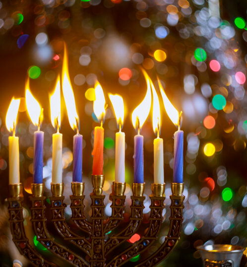 A lit menorah with nine colored candles in gold holders, flames glowing, against a festive multicolored bokeh background.