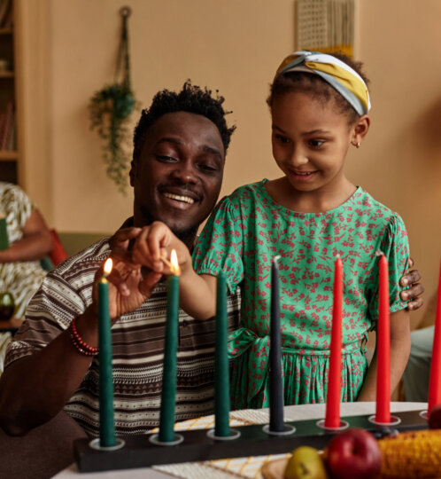 An adult and a young person light candles on a multi-candle holder at a cozy table with fruit nearby, as two others relax in the background.