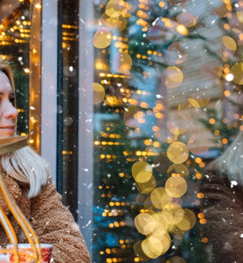 A person with light hair in a brown coat sits by a glass window with warm bokeh lights and falling snow, a candle glowing in a nearby birdcage.