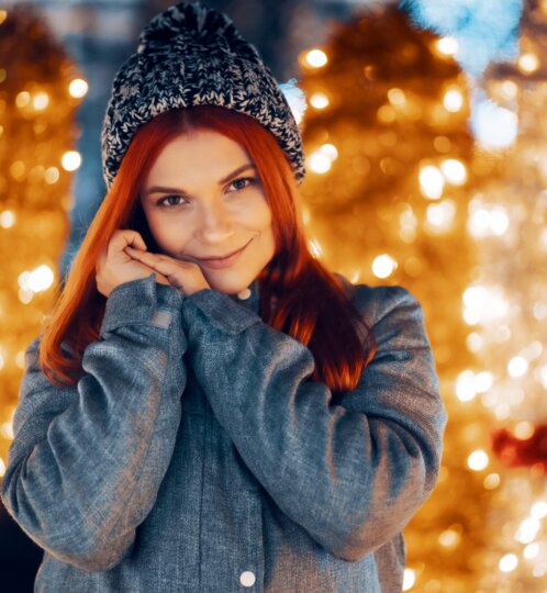 Person with red hair in a knit beanie and denim jacket, smiling with hands at their cheeks, warm golden bokeh lights behind.