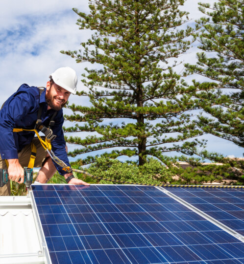 Worker wearing a white hard hat and safety harness kneels on a roof, mounting blue solar panels with a drill.