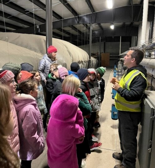 Group of children on a factory tour listening to a guide in a yellow safety vest inside a large industrial facility with ducts and overhead lights.