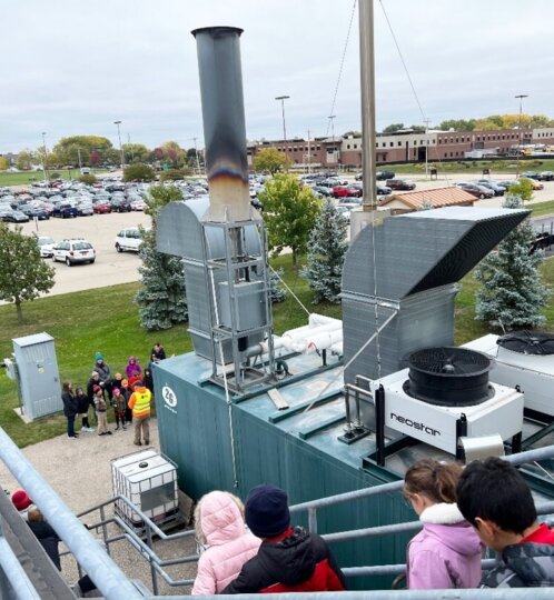 View from a rooftop: industrial HVAC equipment with a tall exhaust stack and a large curved vent, as a group of people in jackets stands at railing.