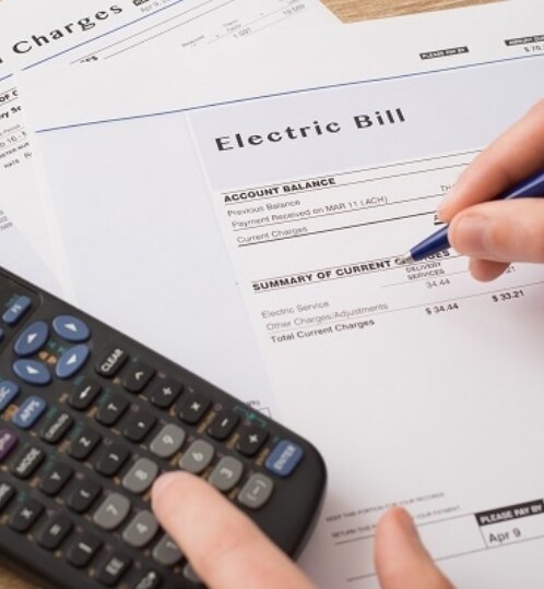 Close-up of a hand holding a blue pen over an electric bill; a calculator and a gas bill sit nearby on a desk.