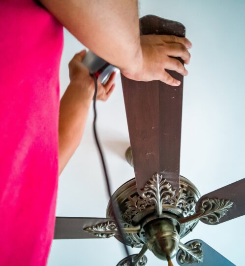 Person in a bright pink shirt holds a power tool against a dark wood ceiling fan blade, with an ornate brass hub visible.