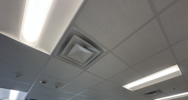 Office ceiling with suspended grid tiles, fluorescent light panels, and a square HVAC vent; windows along the wall show trees and parked cars outside.