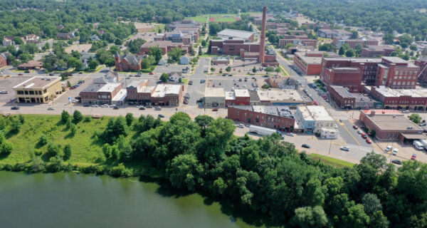 Aerial view of a small town by a wide river; brick buildings along a grid of streets, a tall smokestack, and a tree-lined riverbank.