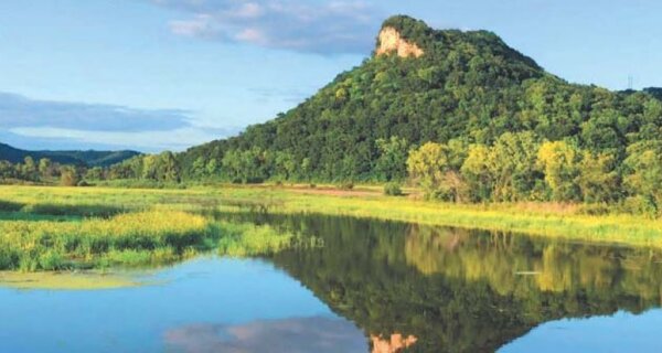 Calm river in a green valley reflects a forested, peak-topped hill and golden trees under a clear blue sky.