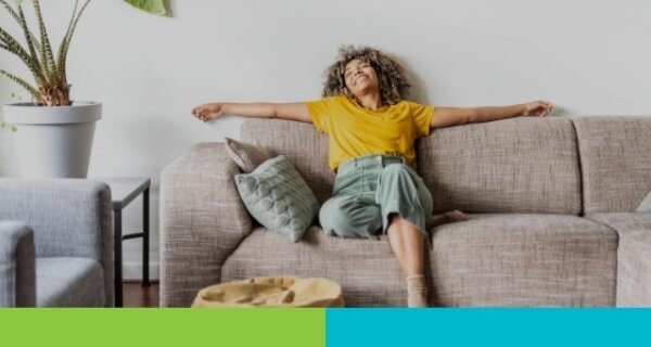 Person with curly hair wearing a yellow shirt and green pants sits on a beige sofa with arms outstretched in a bright living room.