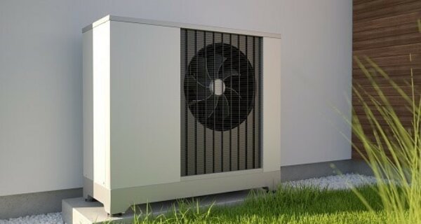 Outdoor air conditioner condenser unit on a small concrete pad beside a white wall, with a visible fan behind a grille, surrounded by gravel and green grass.