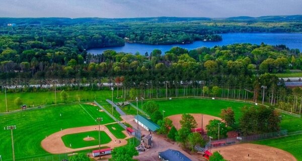 Aerial view of a baseball complex with multiple fields, surrounded by trees, a blue lake beyond, and forested hills on the horizon.