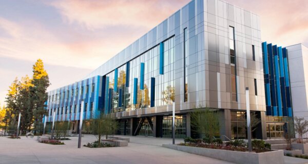 A modern glass building with blue vertical panels stands along a spacious plaza with planters and trees, under a pink-orange sunset sky.